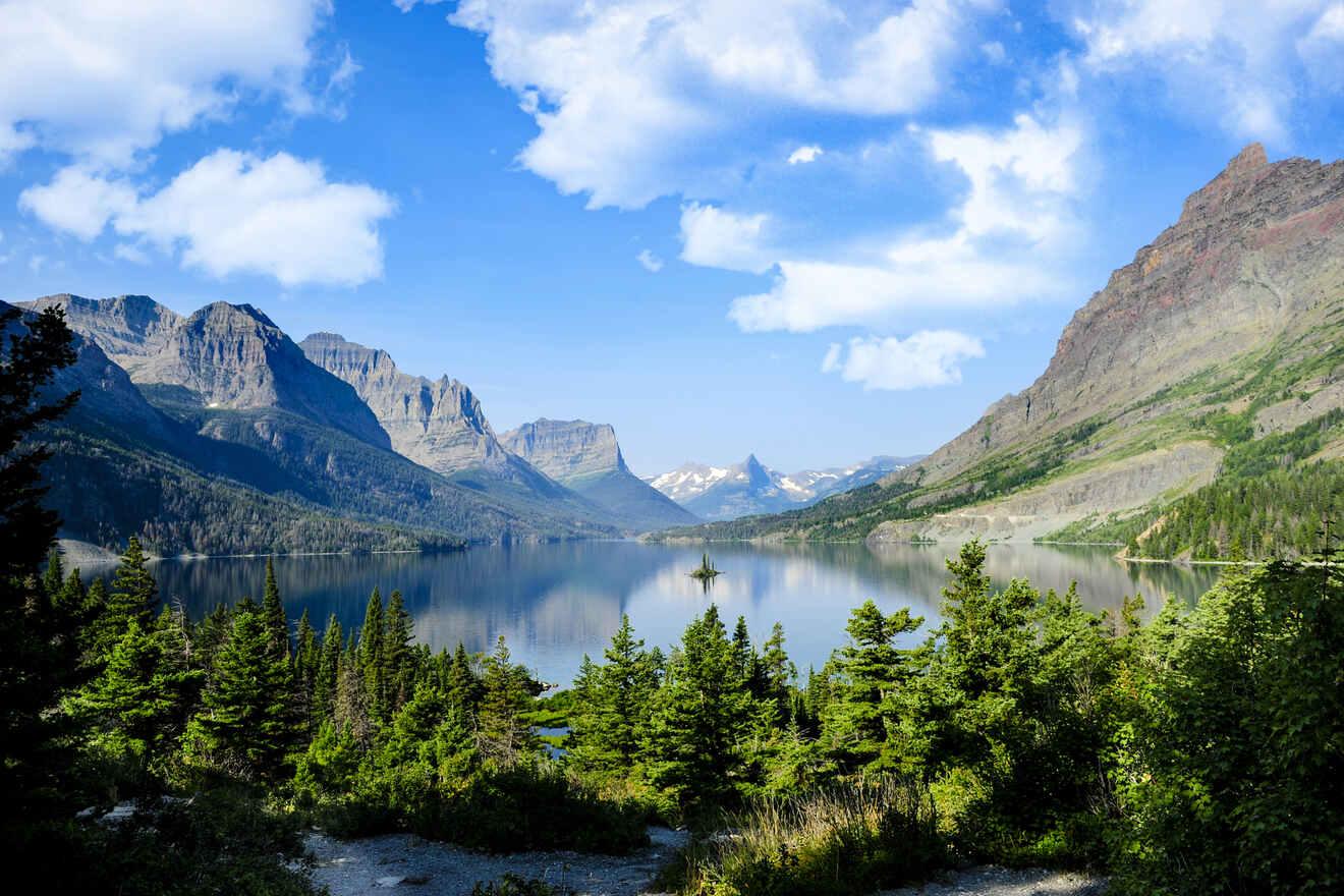 A serene view of Glacier National Park with a clear lake reflecting distant mountains under a blue sky with scattered clouds